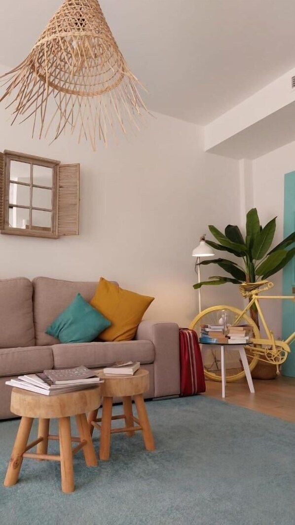 Una sala de estar con un sofá, dos taburetes de madera, una lámpara colgante tejida, una lámpara de pie, una planta en maceta y una bicicleta.