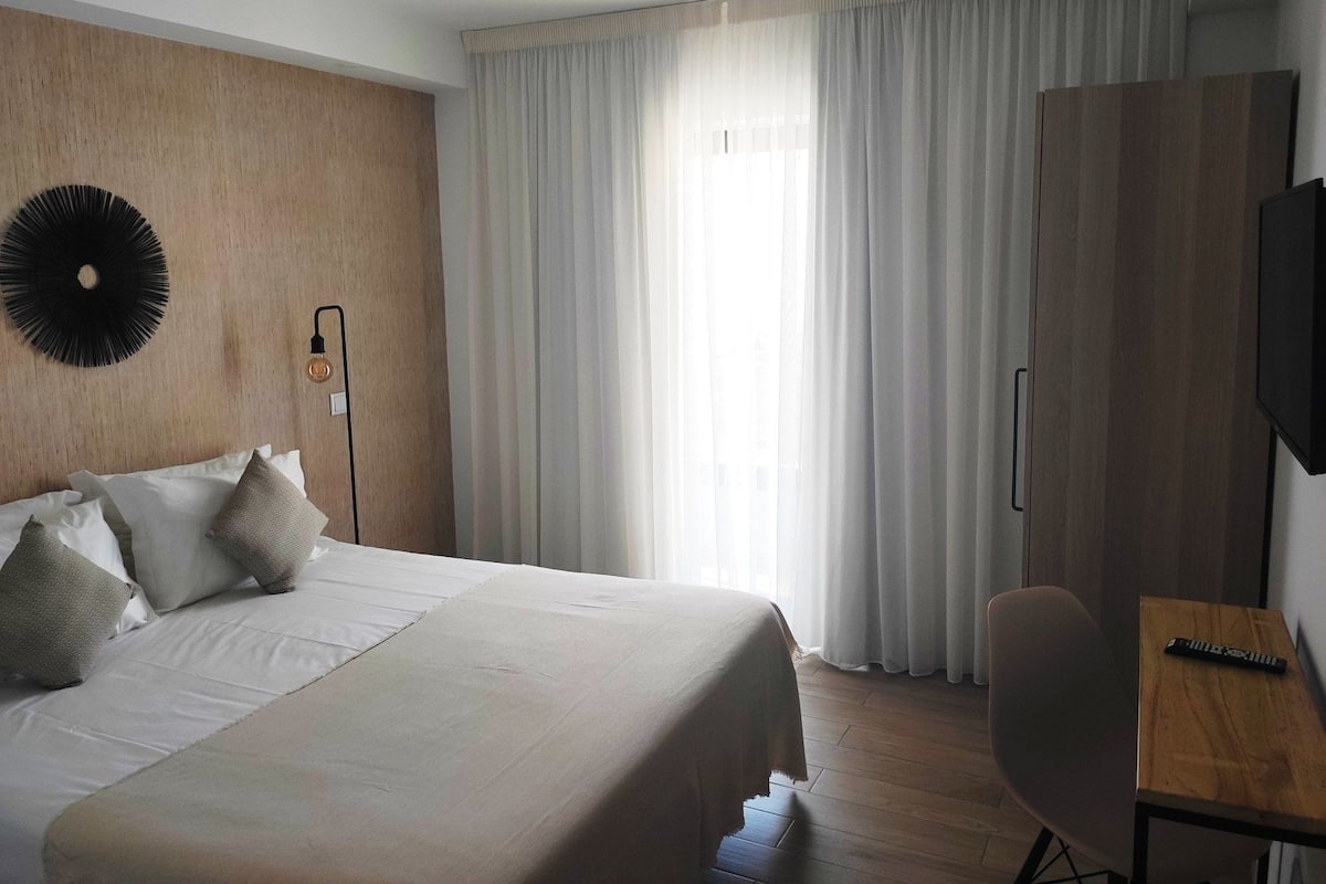 Quarto de hotel com cama grande, escrivaninha, televisão e janela com cortinas.