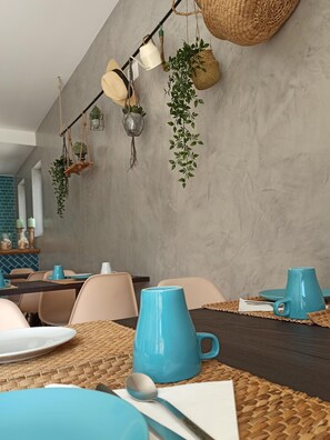 A dining area with a long table set for a meal, featuring woven placemats, blue ceramic mugs, and a variety of hanging plants. - Marsuites B&B (Silves)