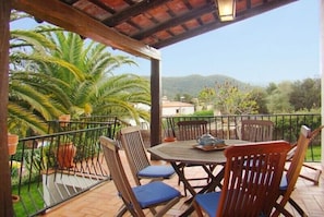 Eine überdachte Terrasse mit Tisch und Stühlen, umgeben von Topfpflanzen und Blick auf Hügel.