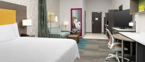 A hotel room with a large bed, a desk with a computer, a chair, a sofa, a mirror, and a television.