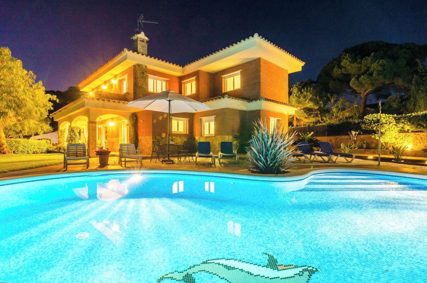 Una casa de dos pisos con piscina, mobiliario exterior y un patio bien iluminado.