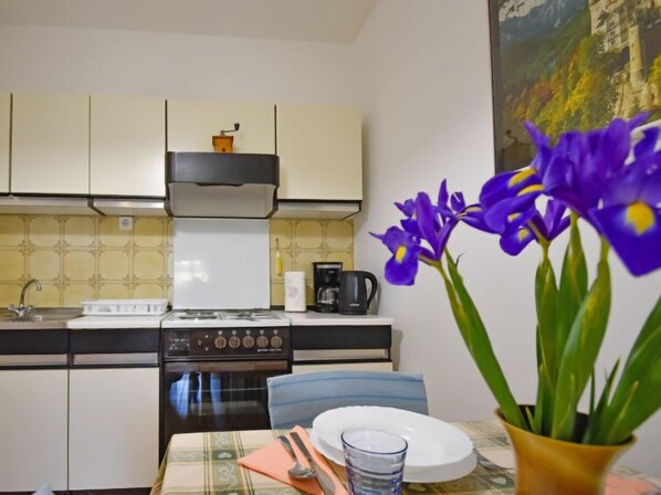 Una cocina con armarios blancos, una estufa y una cafetera. Hay una mesa de comedor con un plato, un vaso y un florero con flores.