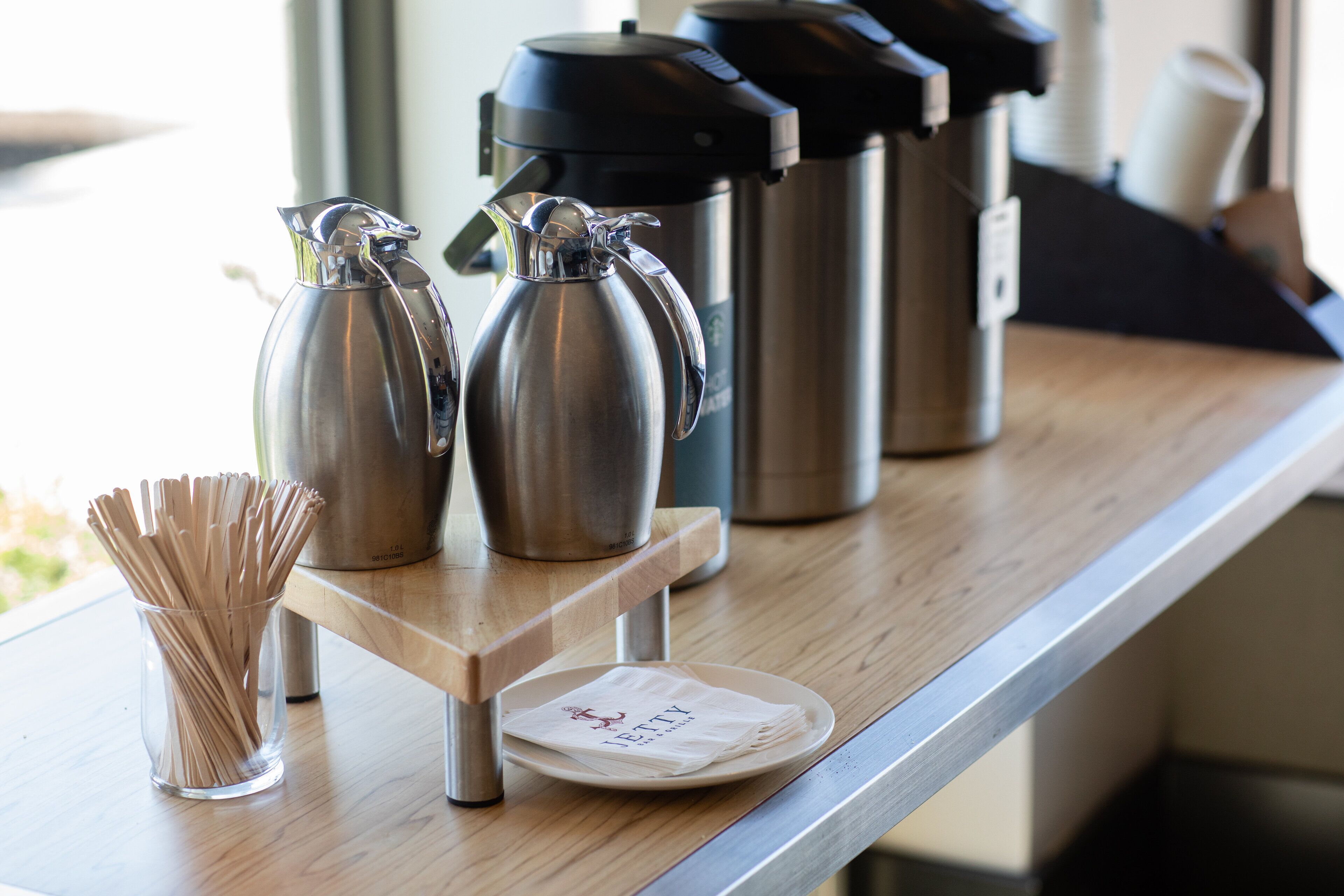 A coffee station with two stainless steel coffee makers, a glass container with straws, and a plate with a napkin on a wooden counter.