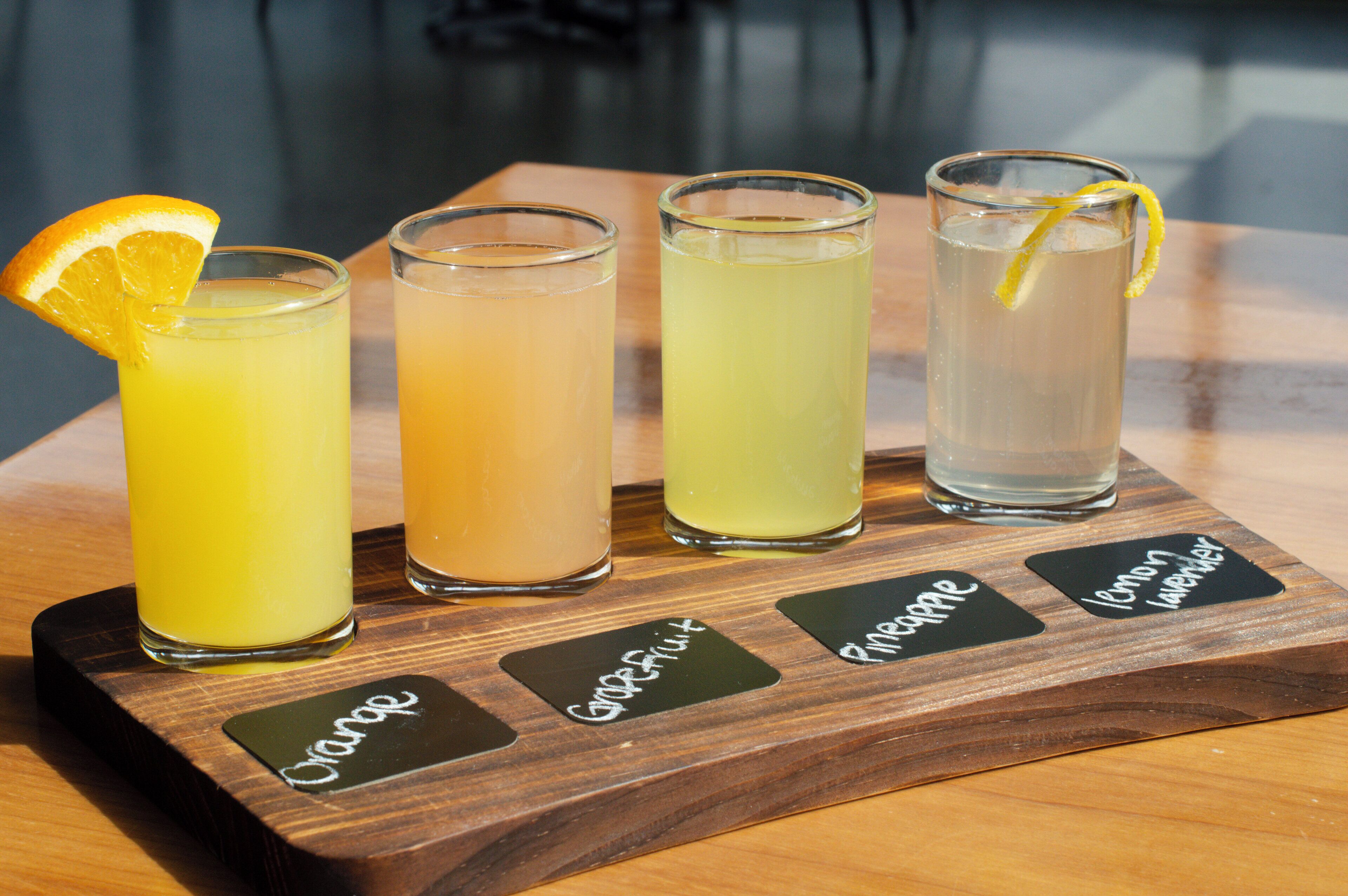 Four glasses of different beverages on a wooden tray with labels.
