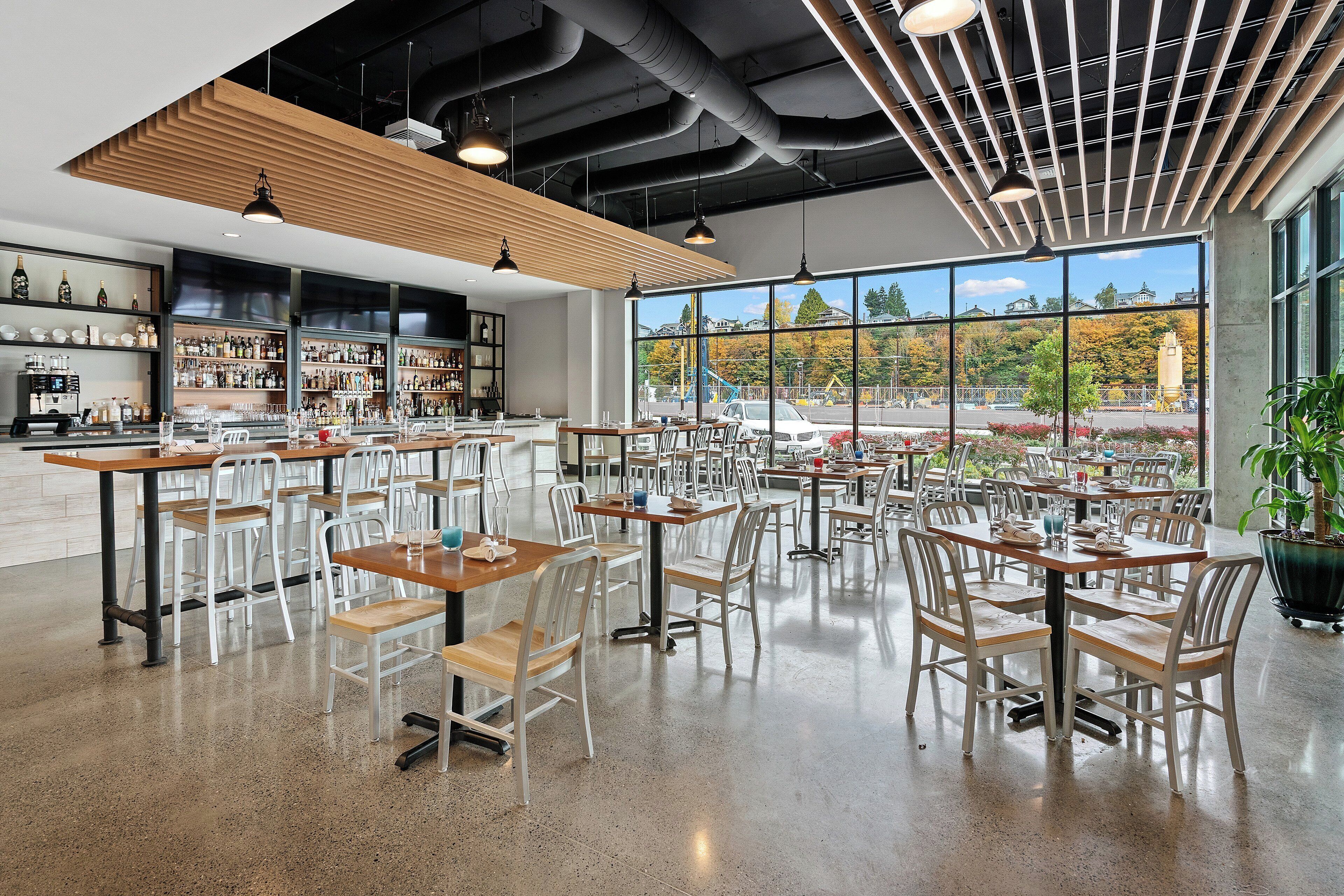 Un amplio comedor interior con varias mesas y sillas, una zona de bar con estantes llenos de botellas, y grandes ventanales que ofrecen vistas al exterior.