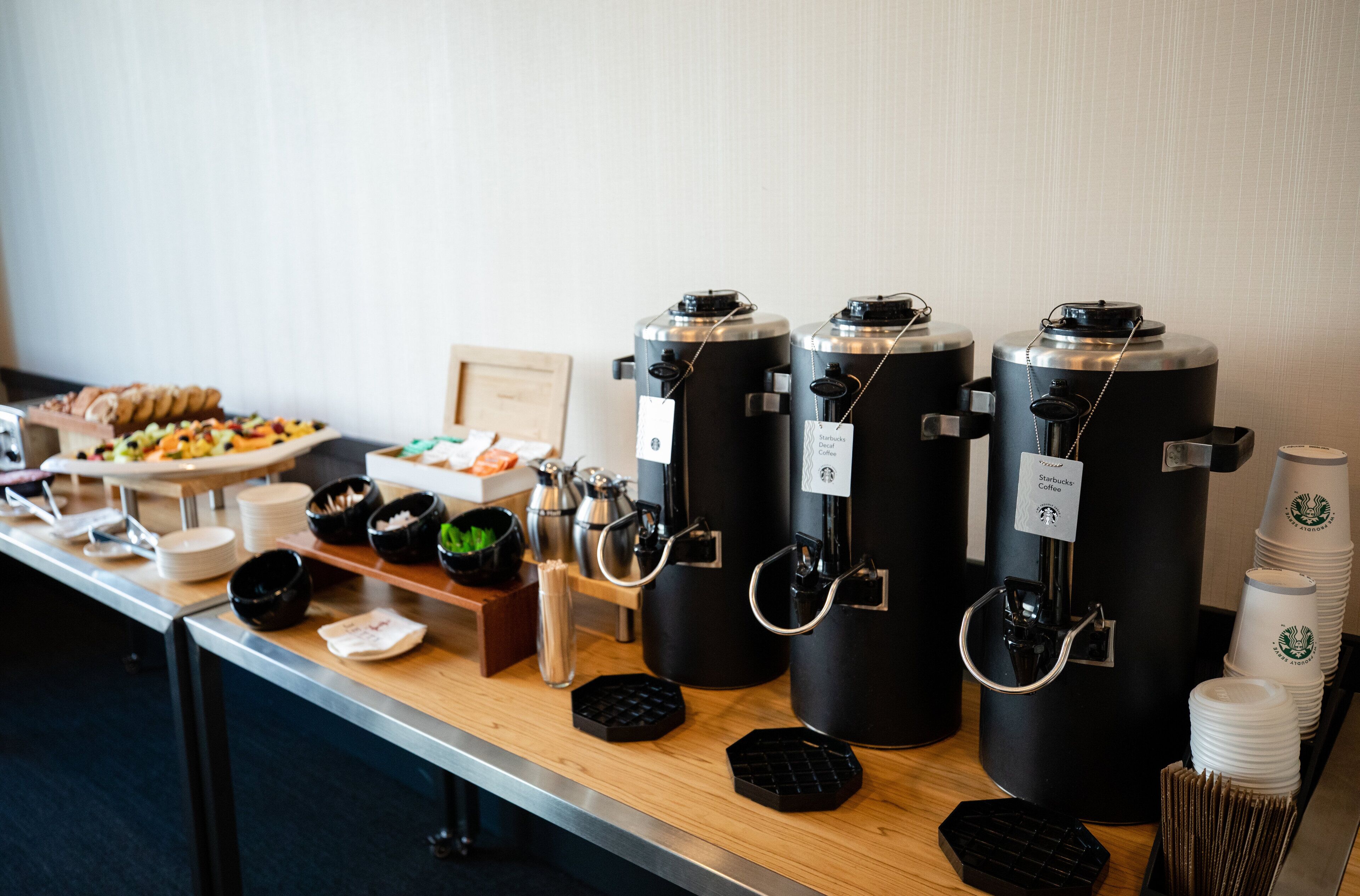 Una barra de desayuno con dispensadores de café, platos, tazones y una bandeja de pasteles.