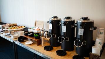 Una barra de desayuno con dispensadores de café, platos, tazones y una bandeja de pasteles.