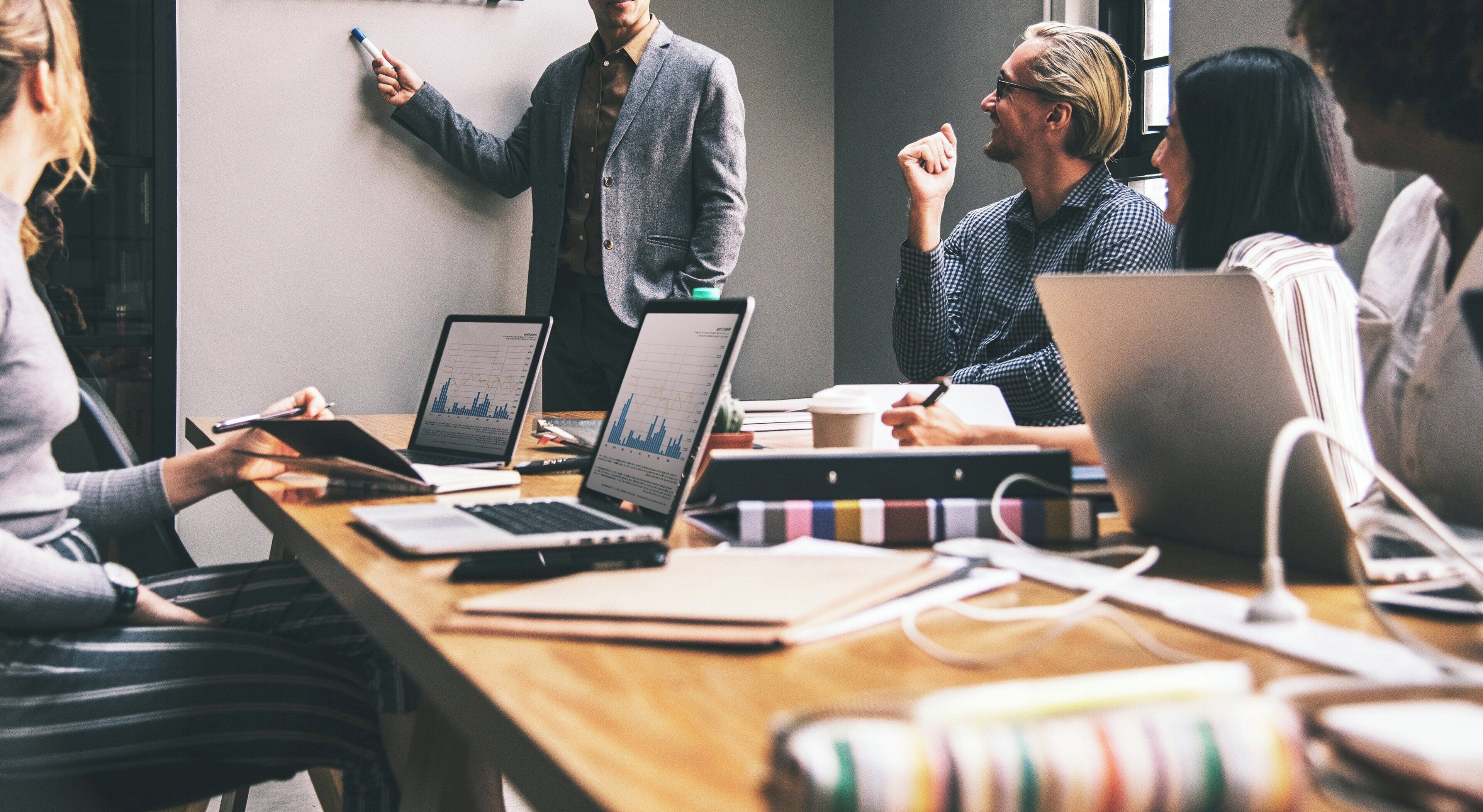 A group of people in a meeting room with laptops, a whiteboard, and a coffee cup on the table.