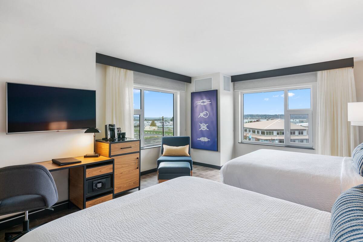 A hotel room with two beds, a desk with a computer, a chair, a TV, and large windows with a city view.