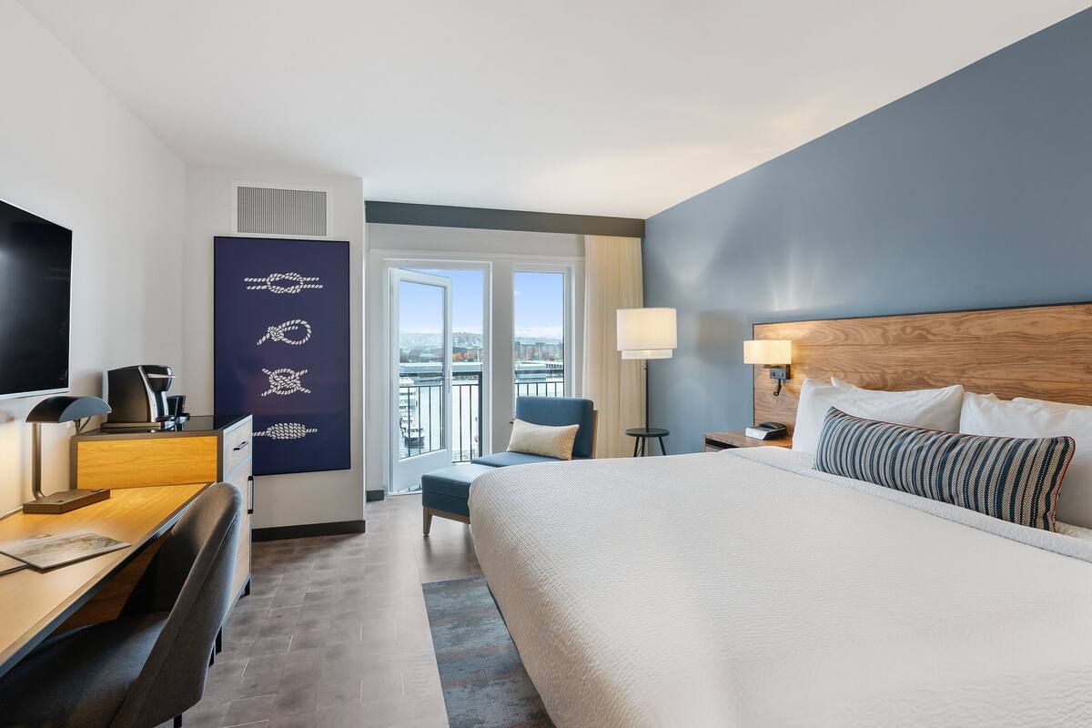 A hotel room with a large bed, a desk with a computer, a chair, a TV, and a view of the city through the windows.