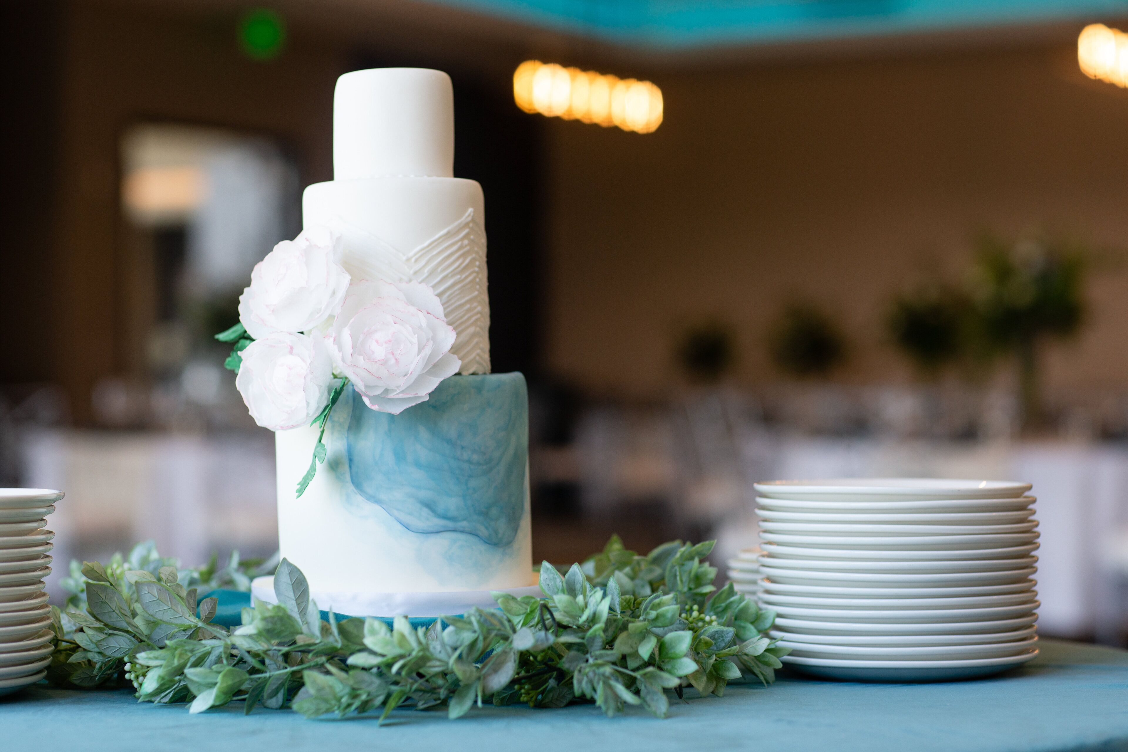 Un pastel blanco de varios niveles con decoraciones florales sobre una mesa con platos blancos y follaje verde.