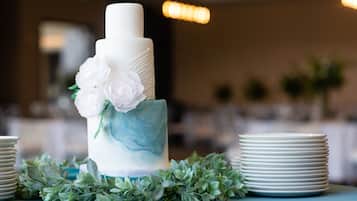 Un pastel blanco de varios niveles con decoraciones florales sobre una mesa con platos blancos y follaje verde.