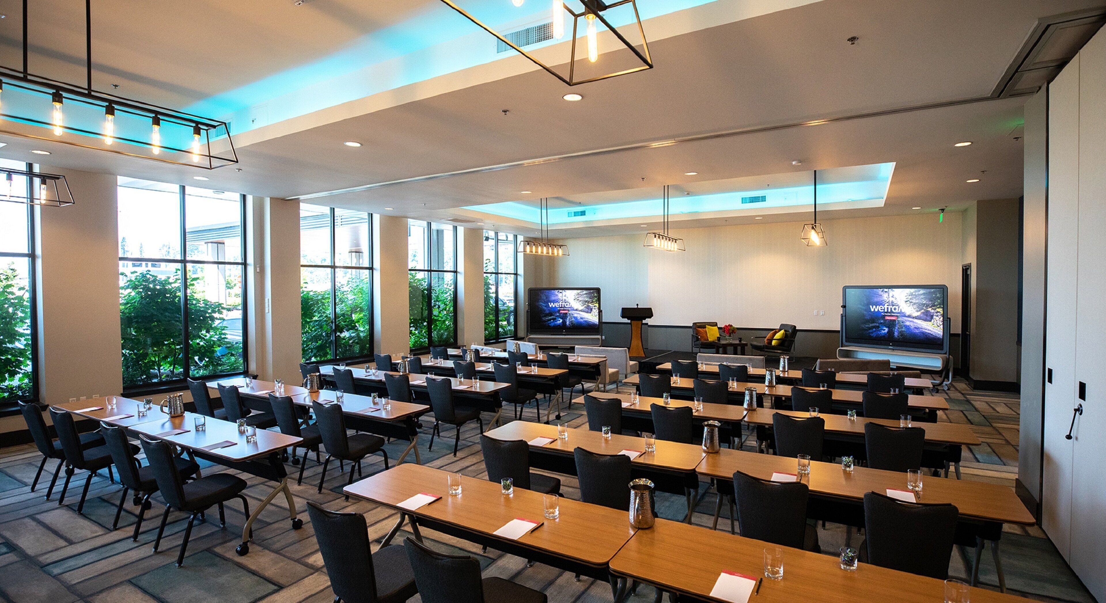 Un salón de conferencias con varias mesas y sillas, ventanas amplias y una pantalla de televisión.