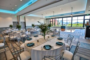 Un amplio salón de banquetes con mesas redondas, sillas blancas y grandes ventanales con vistas a zonas verdes.