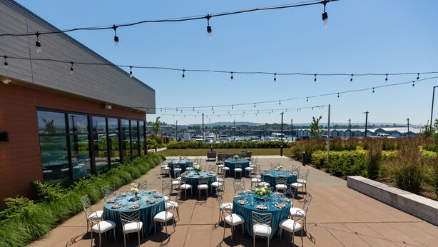 An outdoor patio with round tables and chairs, string lights, and a view of a distant landscape. - Hotel Indigo Everett - Waterfront Place by IHG (Everett)