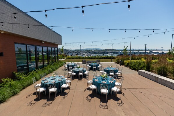 An outdoor patio with round tables and chairs, string lights, and a view of a distant landscape. - Hotel Indigo Everett - Waterfront Place by IHG (Everett)