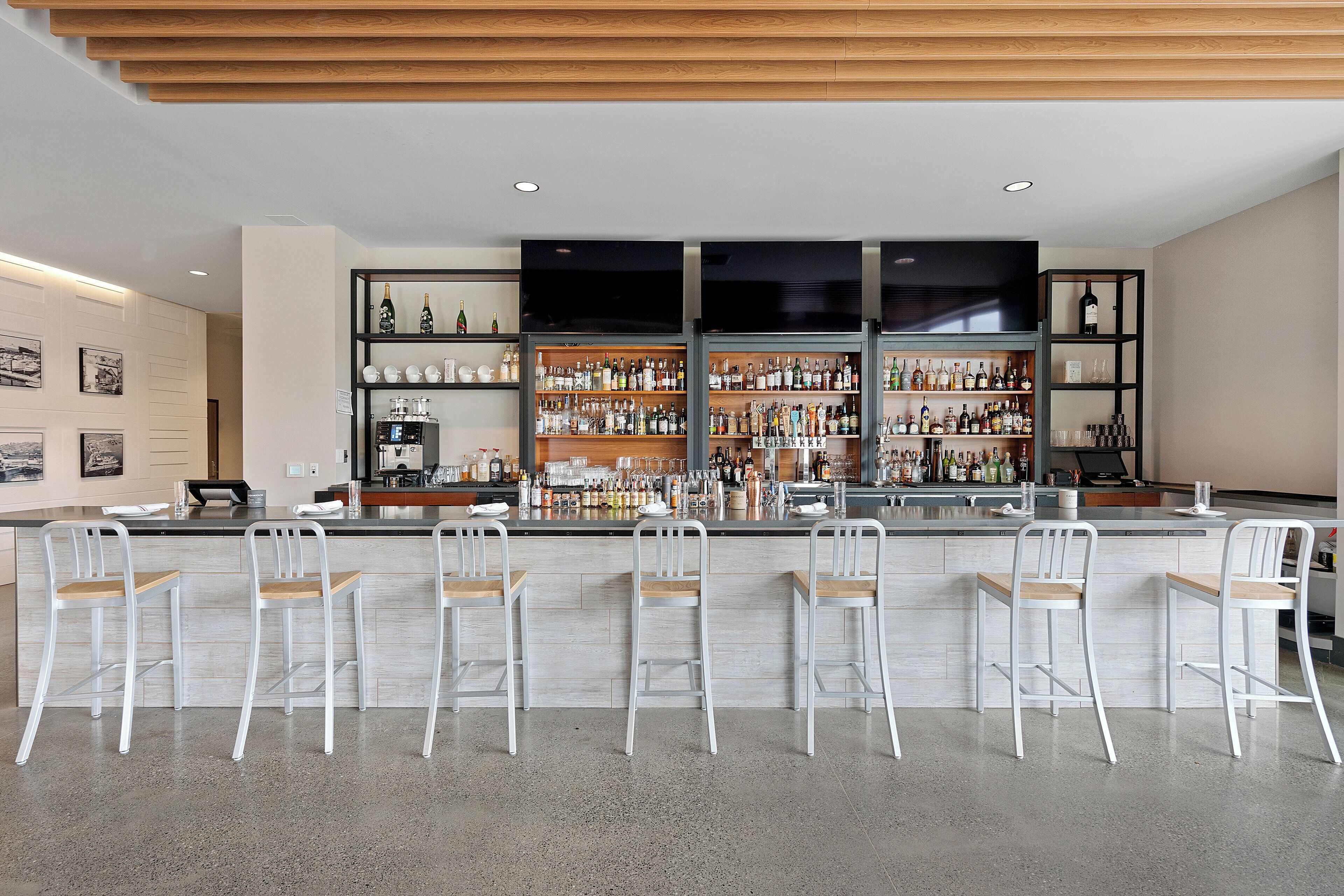 A bar area with a long counter, multiple shelves stocked with bottles, and several white bar stools.