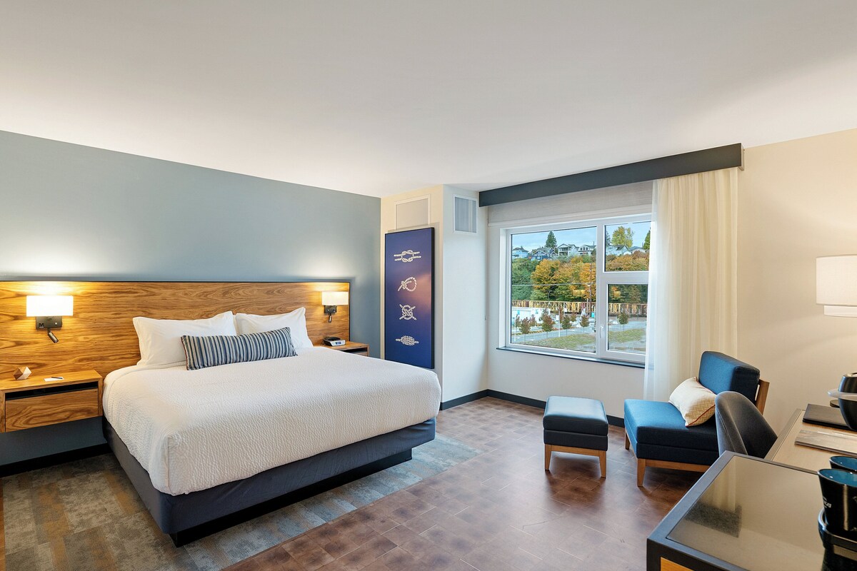 A modern hotel room with a large bed, a desk, a chair, and a view of the outdoors.