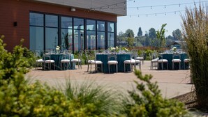 An outdoor patio with tables and chairs, a building with large windows, and string lights. - Hotel Indigo Everett - Waterfront Place by IHG (Everett)