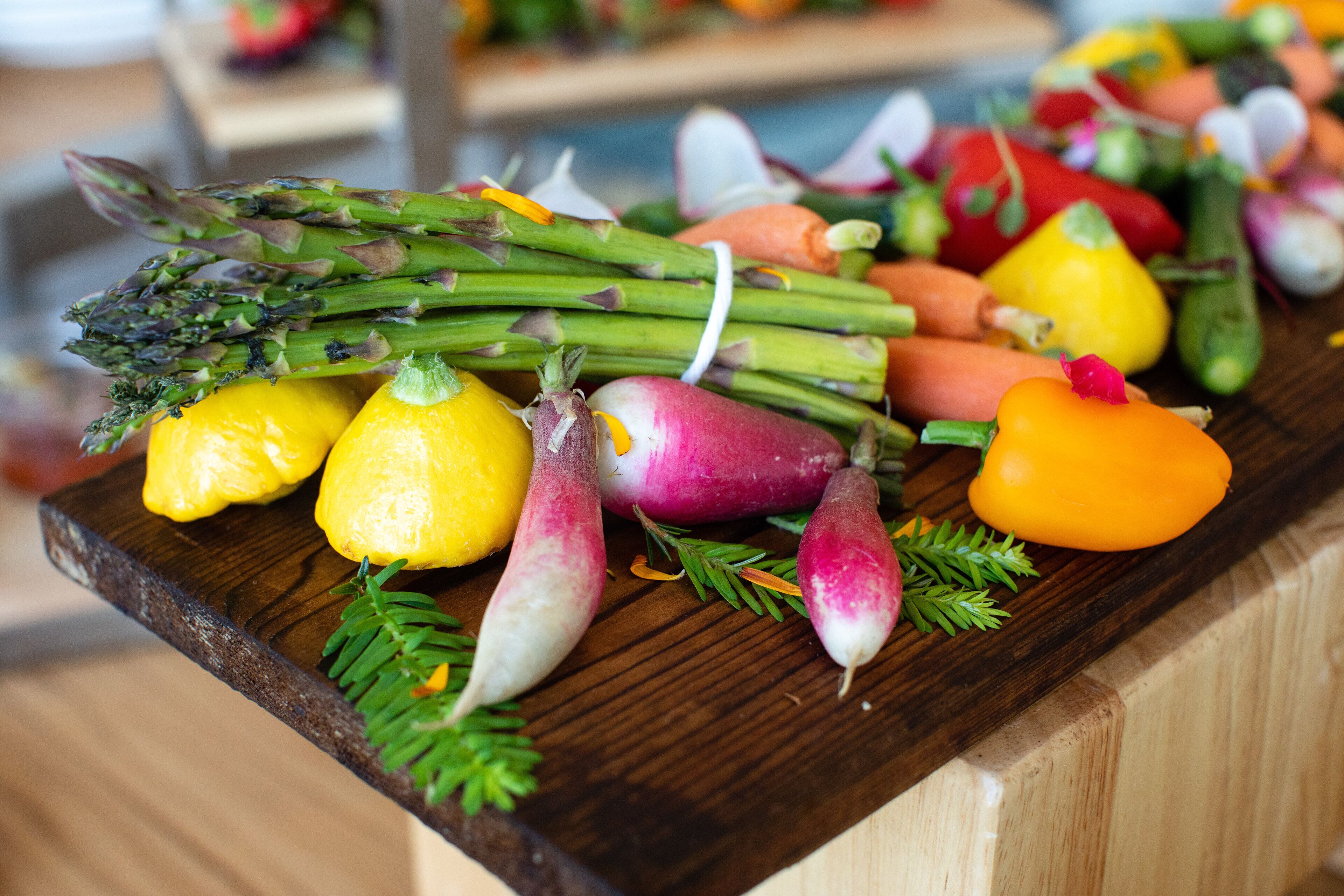 A variety of fresh vegetables including asparagus, lemons, carrots, and radishes on a wooden cutting board.