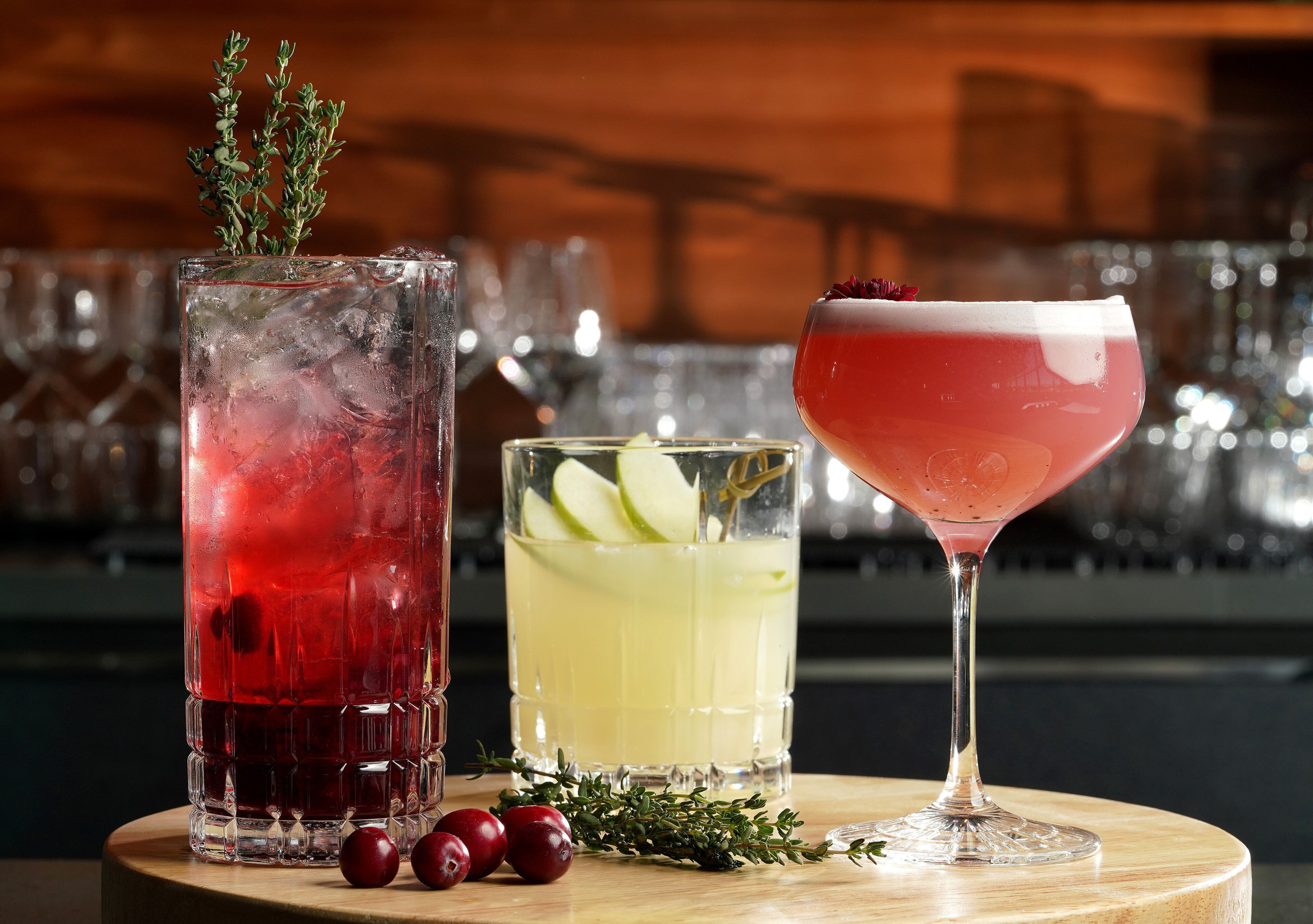 Three glasses of beverages on a wooden table with a blurred background of bar settings.