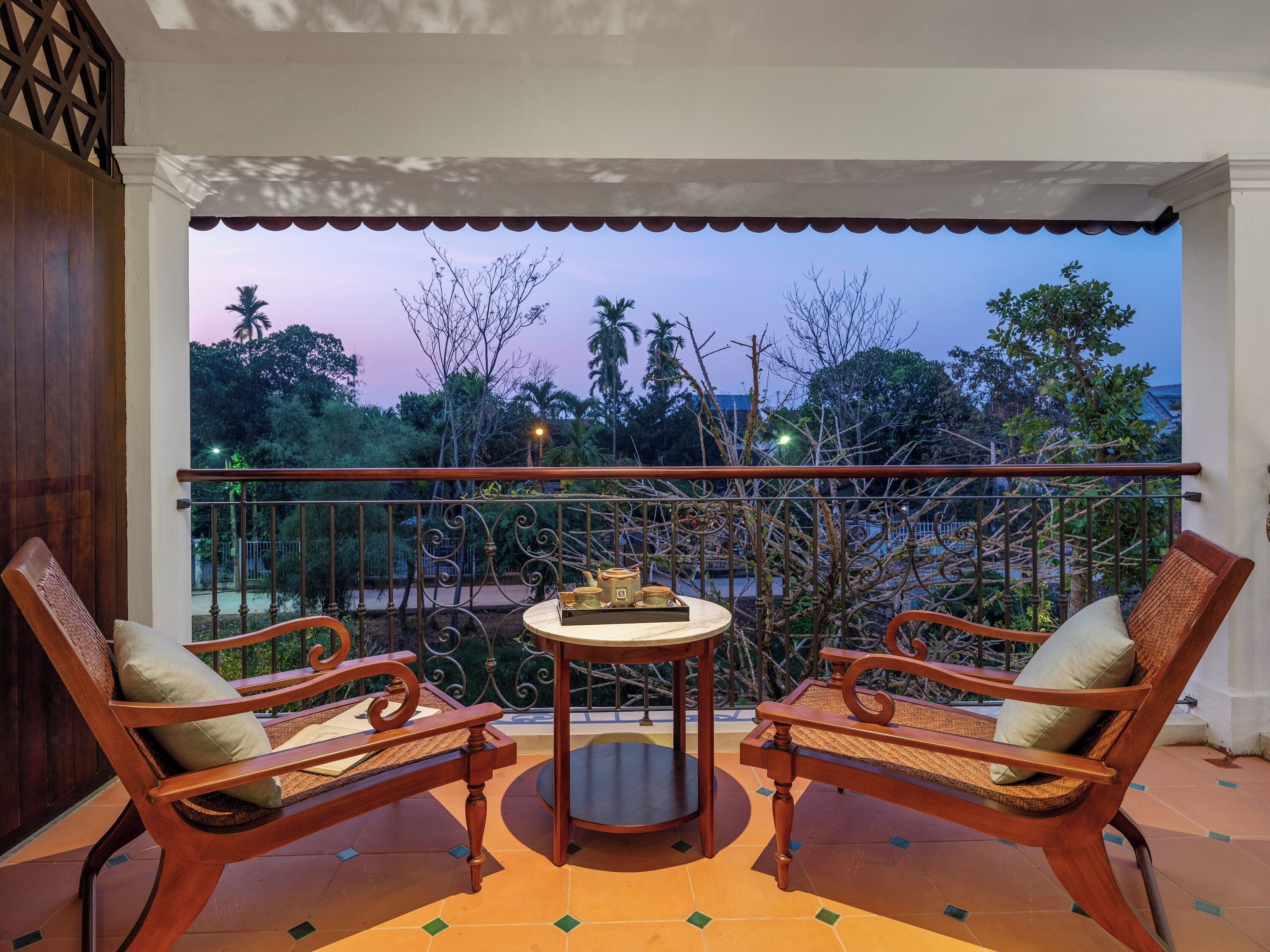 Un balcone con due sedie di legno e un tavolino, affacciato su un giardino con alberi e un cielo sereno.
