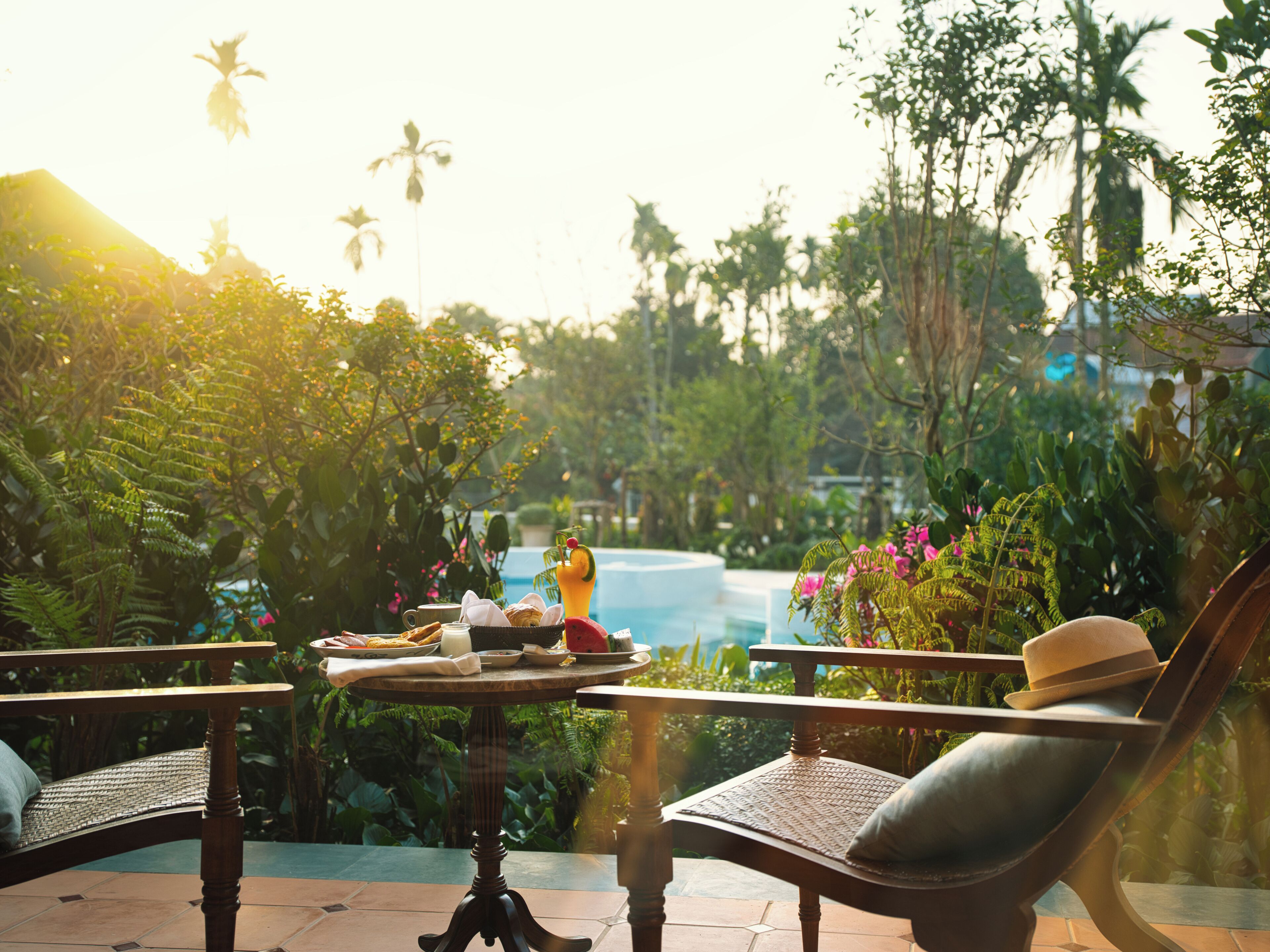 A patio with a round table set for breakfast, surrounded by lush greenery and a pool in the background.