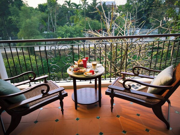A balcony with two wooden chairs, a small round table with food and drinks, and a view of greenery and trees.