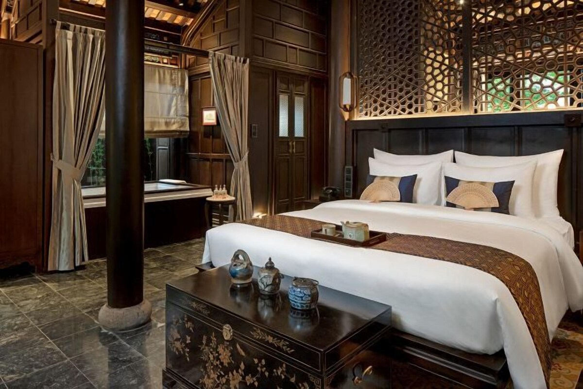 A bedroom with a large bed, a nightstand, a decorative chest, and a window with curtains.