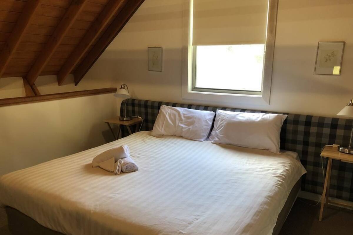 A neatly made bed with white linens and pillows, a window with blinds, a small bedside table with a lamp, and a checkered wall panel.