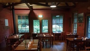 A restaurant with wooden tables and chairs, large windows with a view of greenery, and a rustic interior with exposed beams. - Corinna Wilderness Village (Corinna)