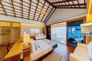 A spacious bedroom with a wooden ceiling, a large bed, a desk, and a view of the ocean through a sliding glass door.