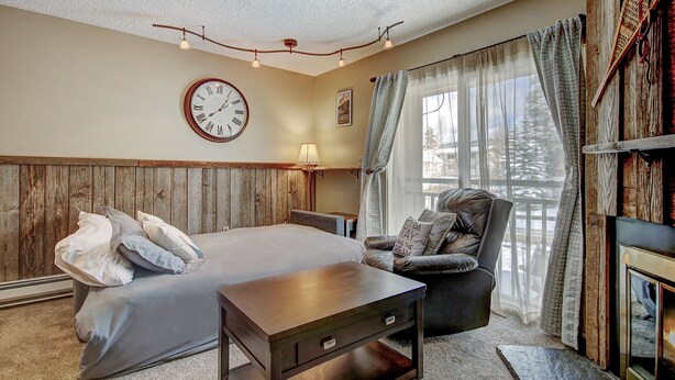 A cozy bedroom with a wooden headboard, a bed with pillows, a wooden nightstand, a chair, a clock, and a window with curtains.