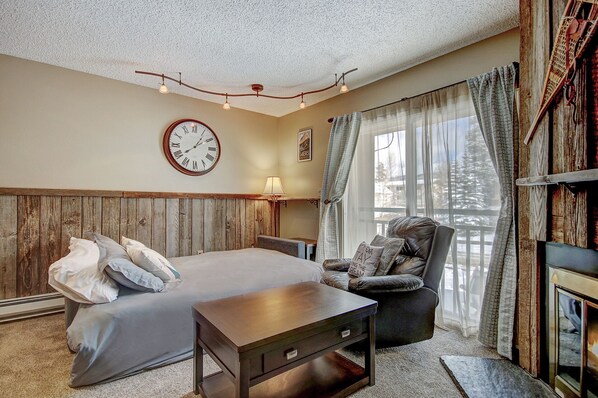 A cozy bedroom with a wooden headboard, a bed with pillows, a wooden nightstand, a chair, a clock, and a window with curtains.