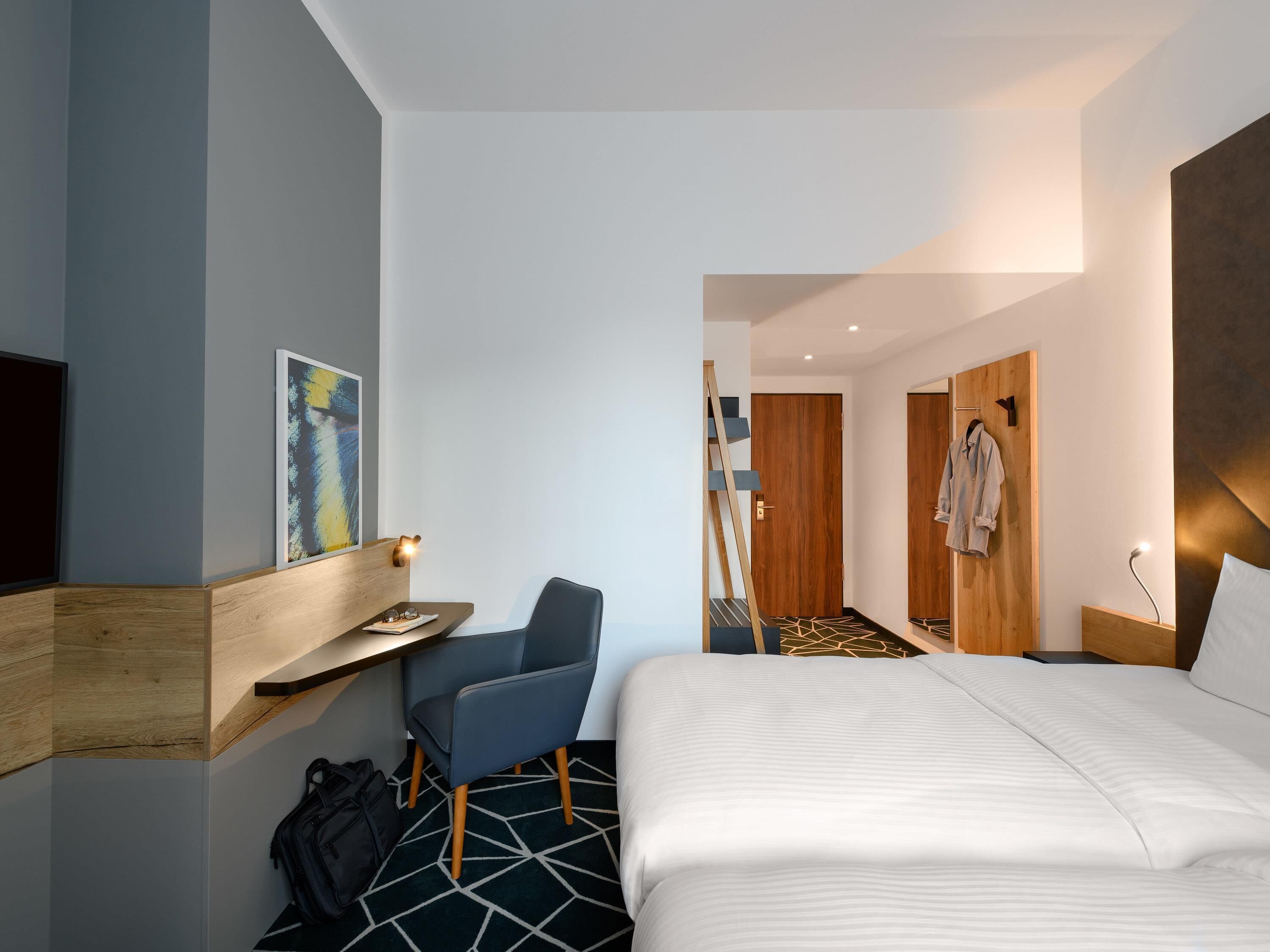 Una habitación de hotel moderna con una cama, un escritorio, una silla, un perchero y un armario de madera.