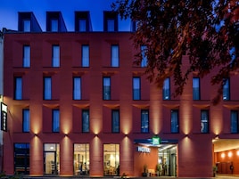 Ein modernes Hotelgebäude mit roter Fassade, zahlreichen Fenstern und einem gut erkennbaren Eingang mit einem Schild.