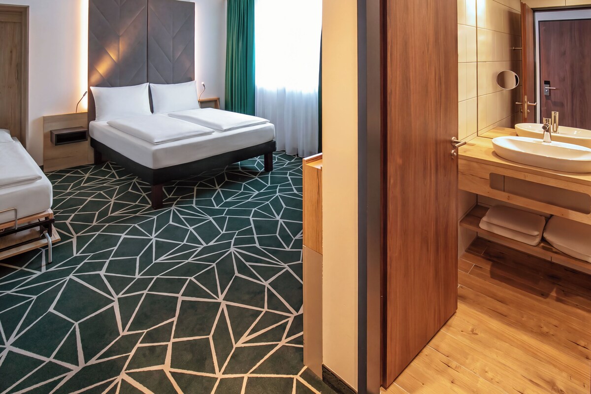 A hotel room with a bed, bedside tables, a bathroom with a sink and mirror, and a geometric patterned carpet.