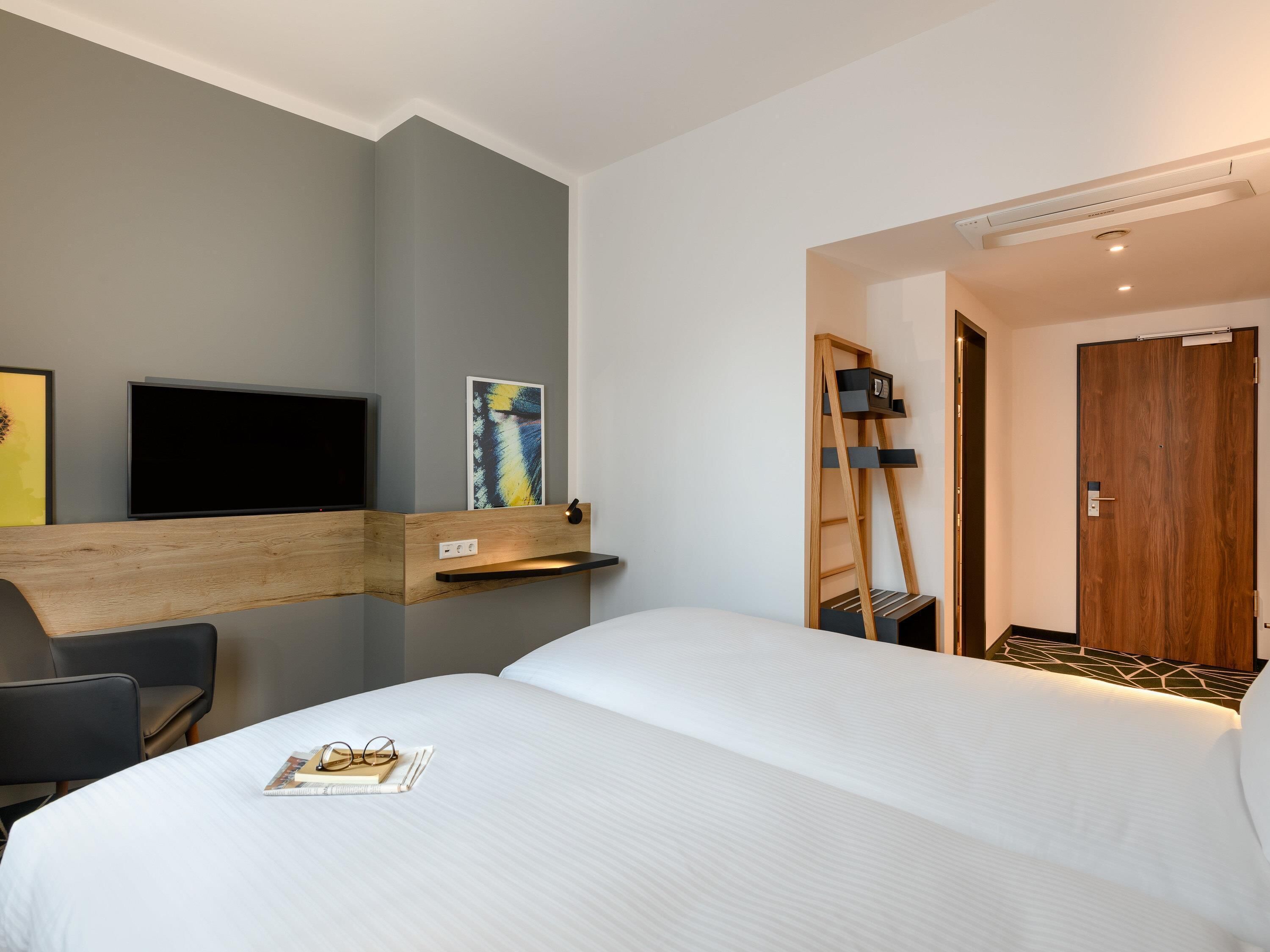 A hotel room with a bed, a desk with a chair, a TV mounted on the wall, and a door.