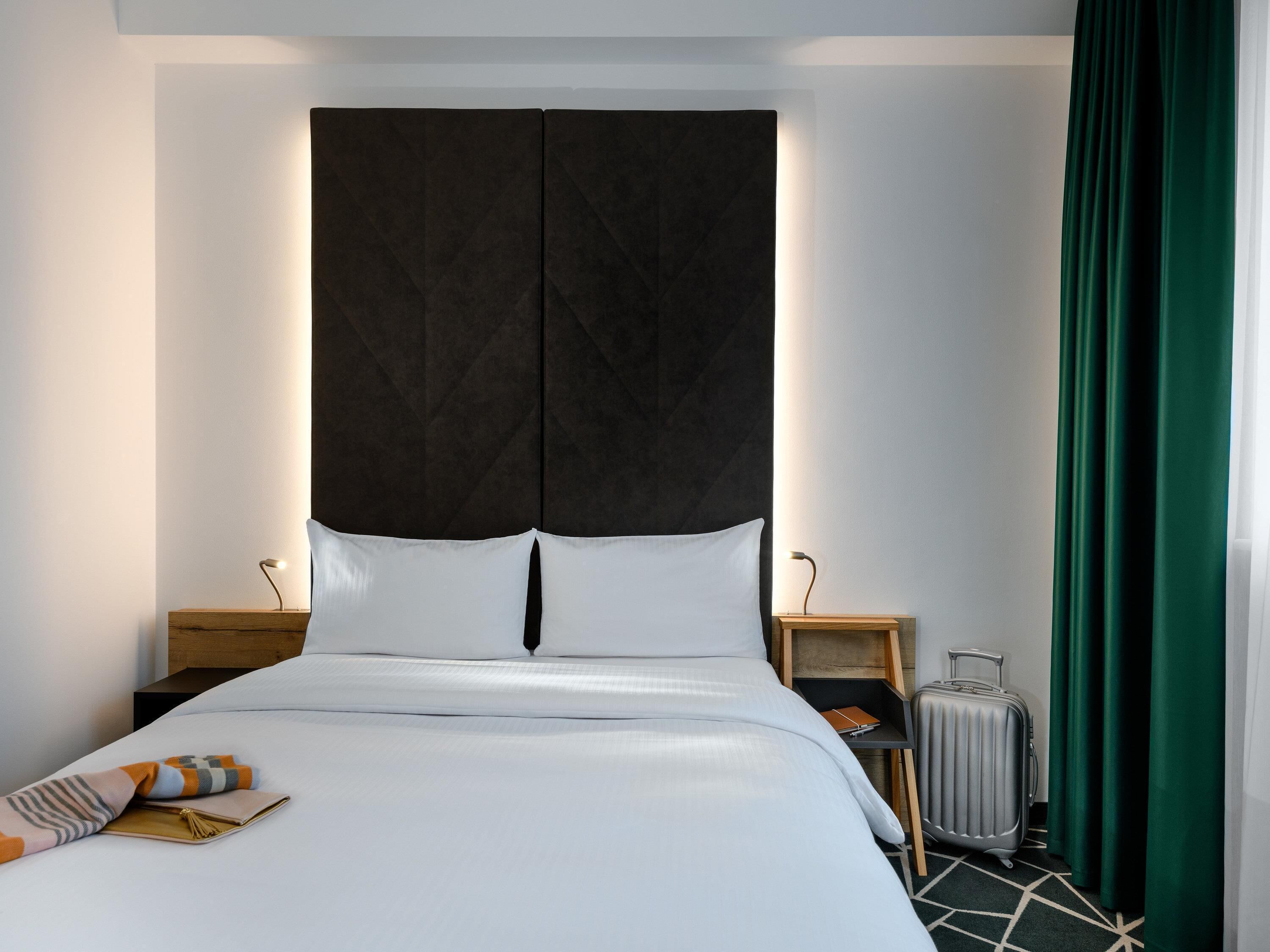 Ein Hotelzimmer mit einem großen Bett, einem dunklen Kopfteil, zwei Nachttischen mit Lampen, einem Koffer und einem grünen Vorhang.