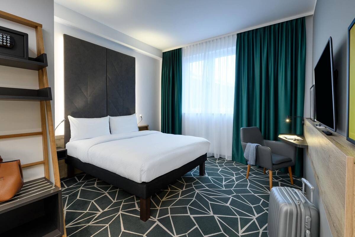A hotel room with a bed, a desk with a chair, a television, and a suitcase.