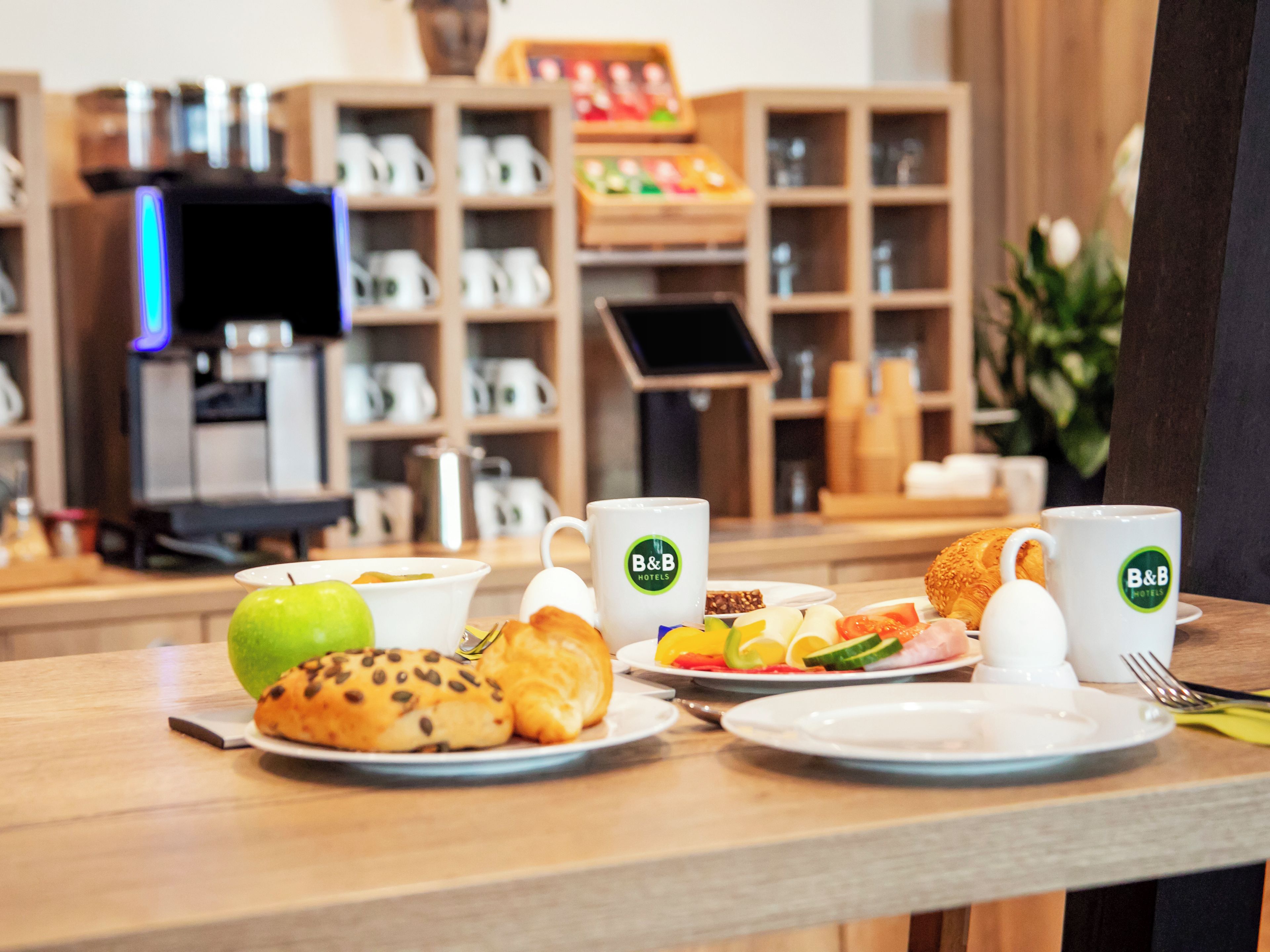A breakfast setup with a variety of food items and B&B hotel branded mugs on a wooden table.