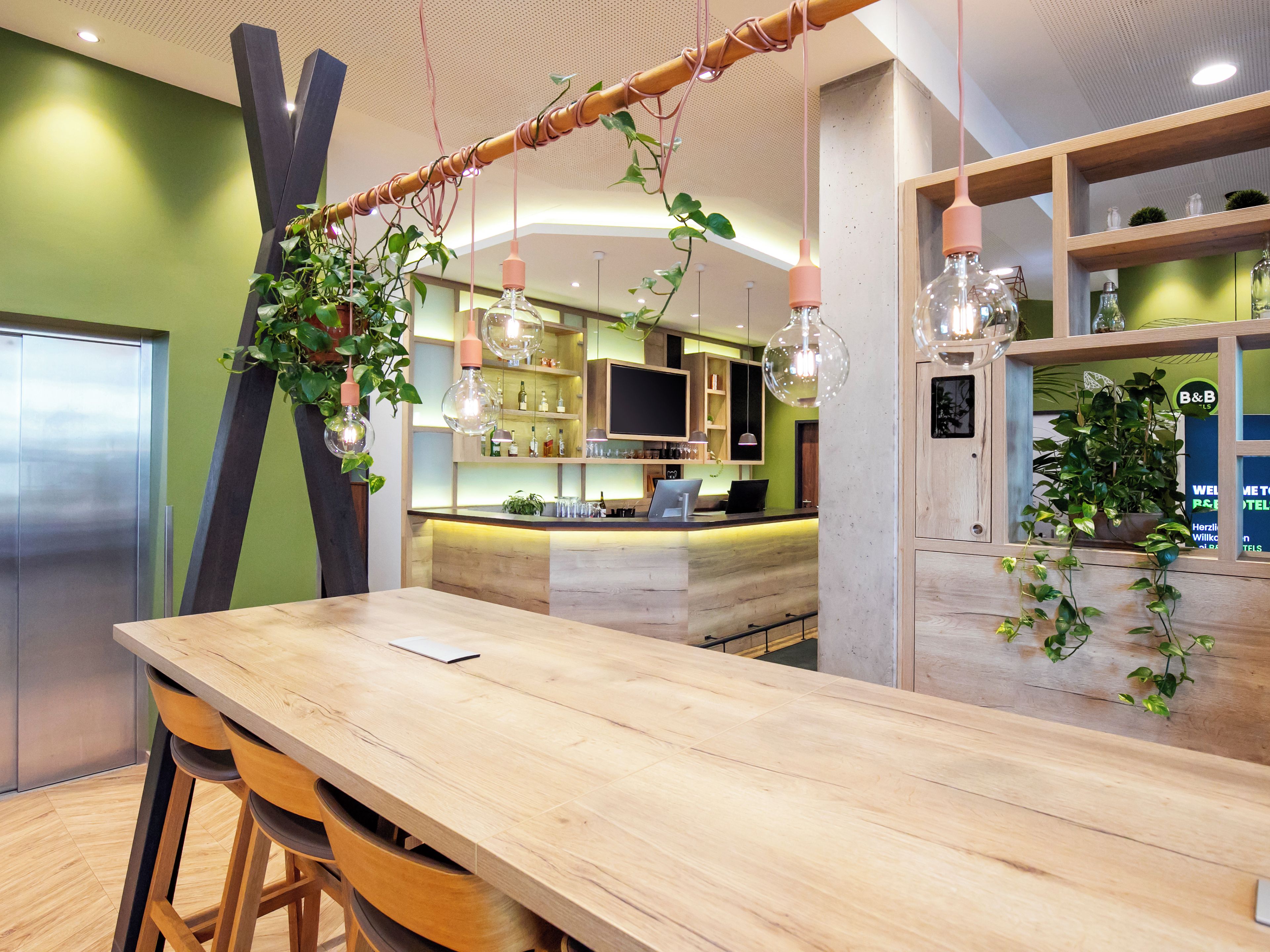 A modern bar area with a wooden counter, bar stools, hanging light fixtures, and green plants.