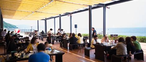 An outdoor dining area with tables and chairs, a covered section, and a view of the sea.