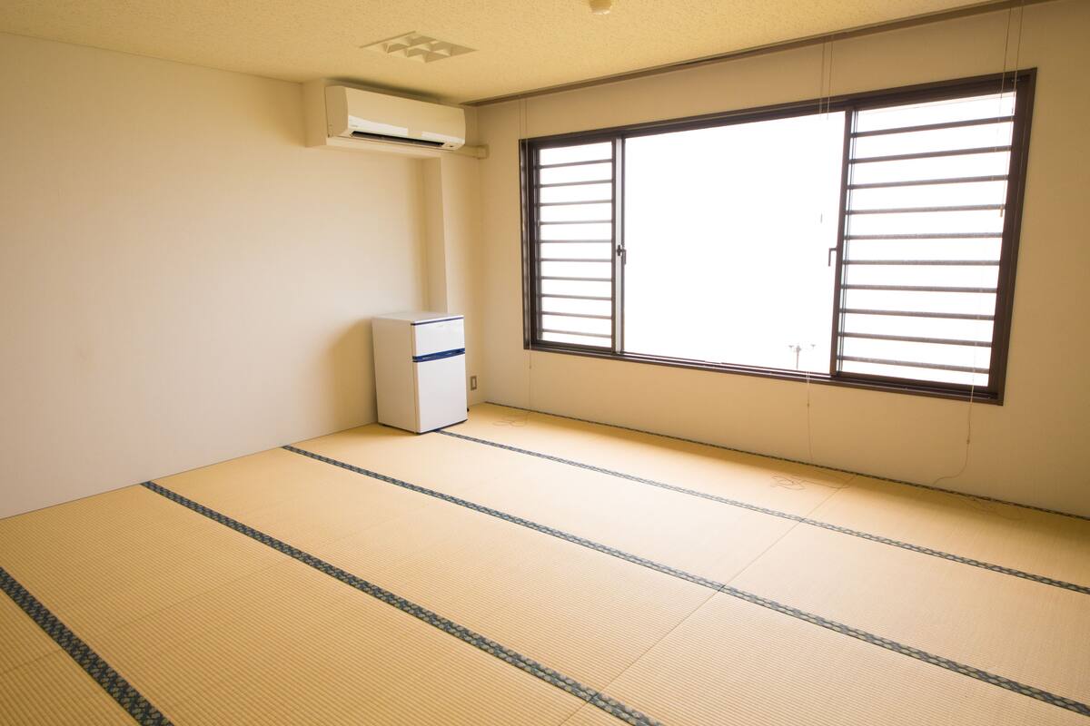 A room with tatami flooring, a large window with blinds, and a small air conditioning unit.