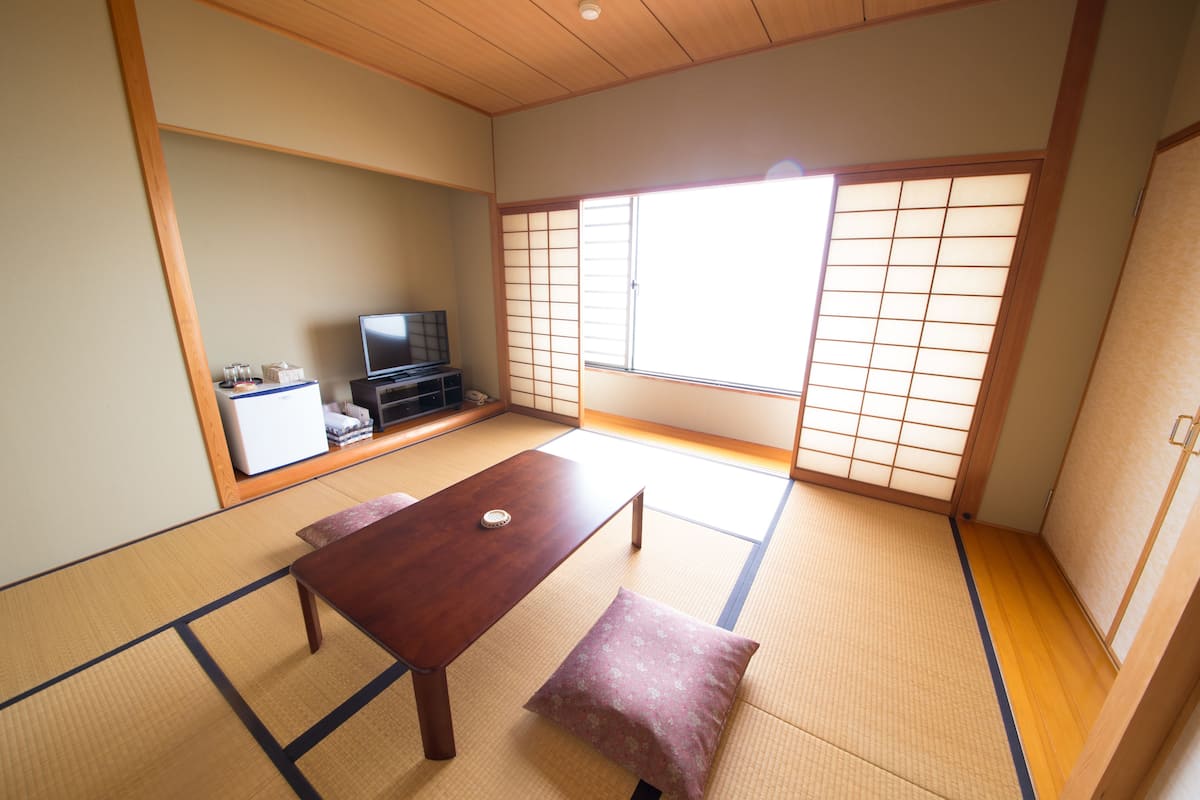 A traditional Japanese room with tatami flooring, a low wooden table, a bench, a television, and sliding doors.