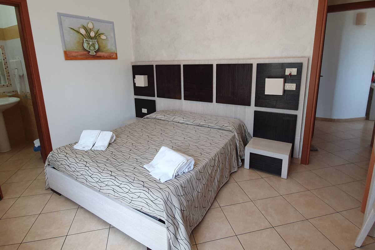 Una cama bien hecha con una colcha estampada y dos toallas dobladas, un aplique en la pared y una mesita de noche pequeña.