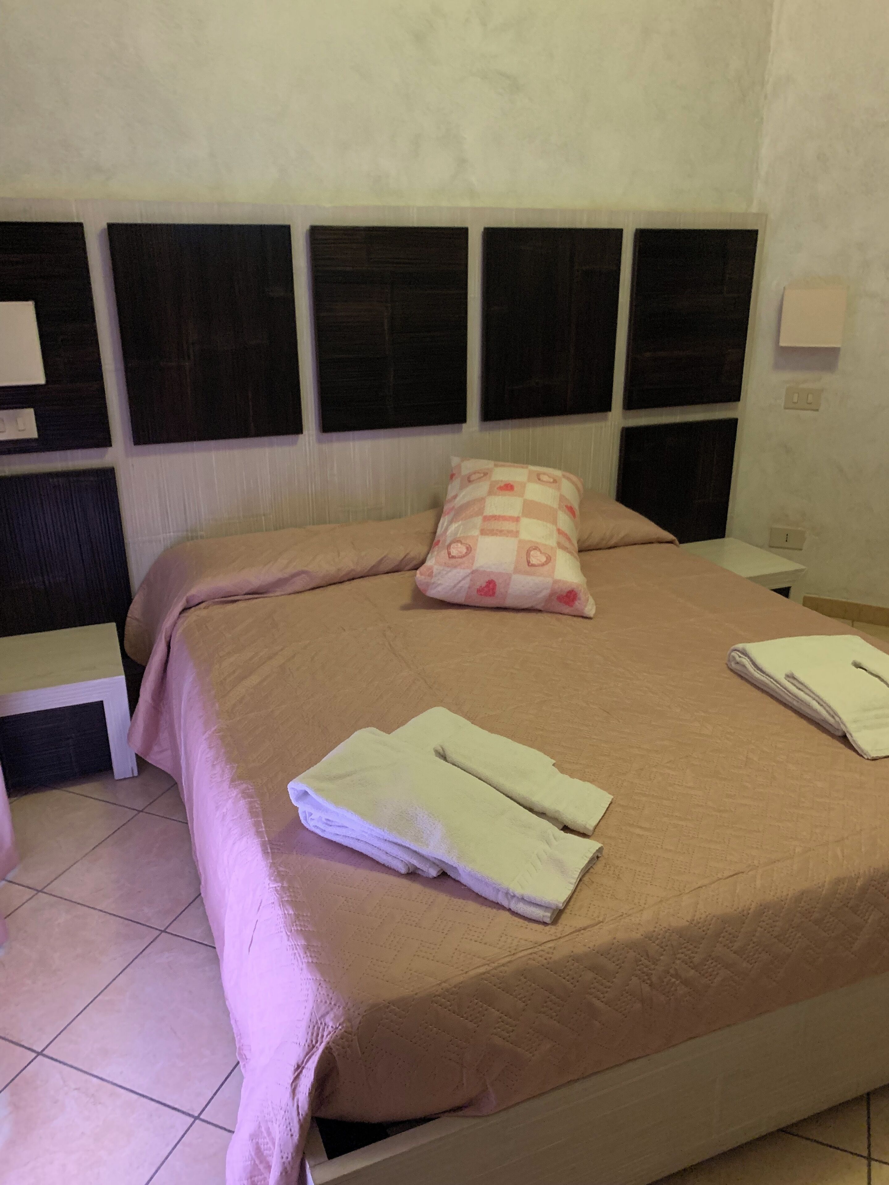 A neatly made bed with a beige bedspread and two pillows, a small bedside table with a lamp, and a wall-mounted light fixture.