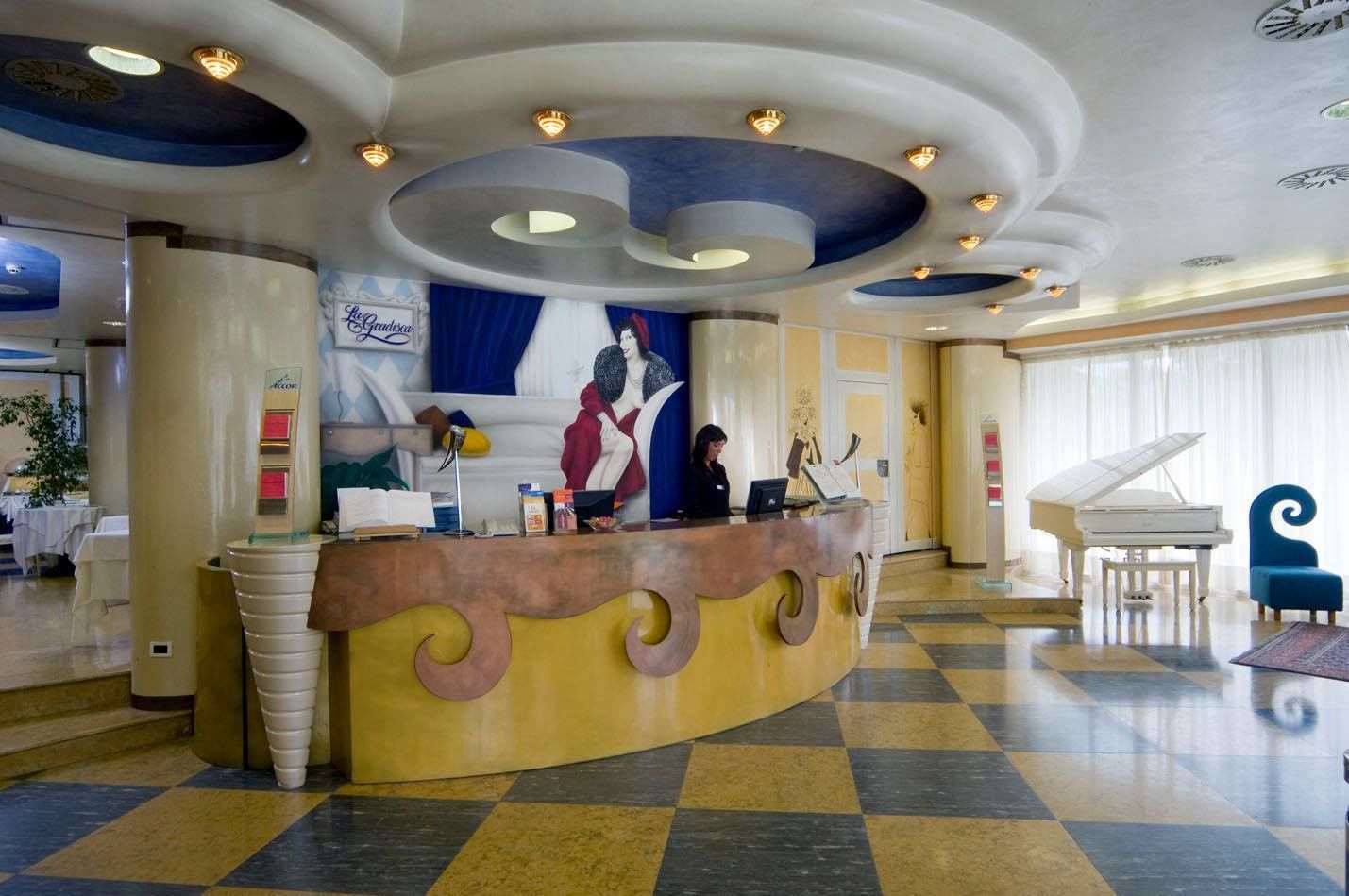 A hotel lobby with a reception desk, a grand piano, and a large mural on the wall.
