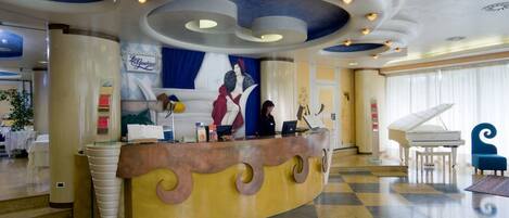 A hotel lobby with a reception desk, a grand piano, and a large mural on the wall.